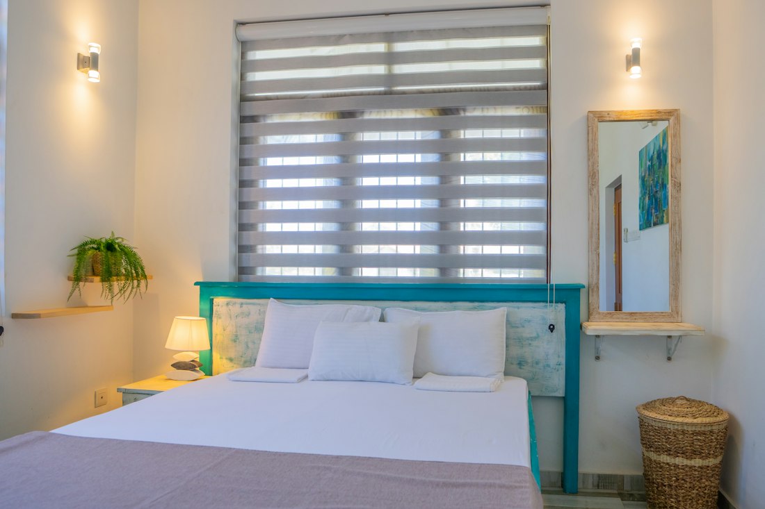 Chambre claire avec un grand lit blanc, tête de lit turquoise, store tamisé et plante verte.