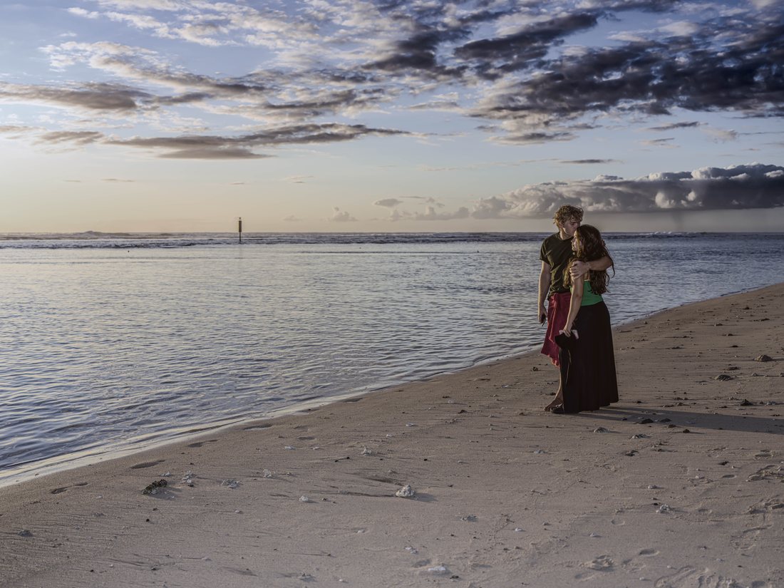 Couple enlacé sur le sable clair au bord du lagon, sous un ciel nuageux de fin de journée.