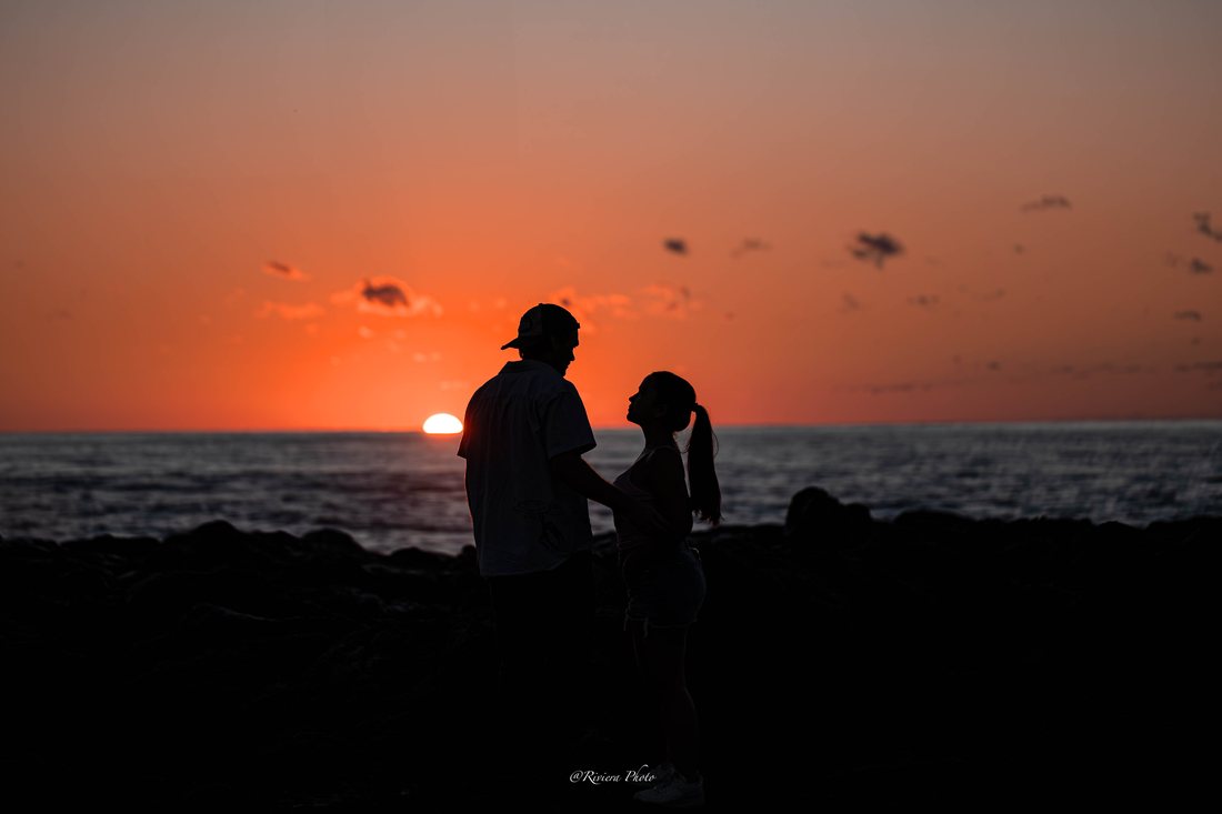 Silhouette d'un couple face à face sur des rochers, devant l'océan, au moment où le soleil touche l'horizon.