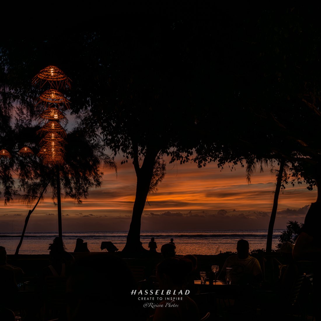 Terrasse au bord de la mer au coucher du soleil, sous les arbres, des lanternes tressées allumées et des convives en silhouette.