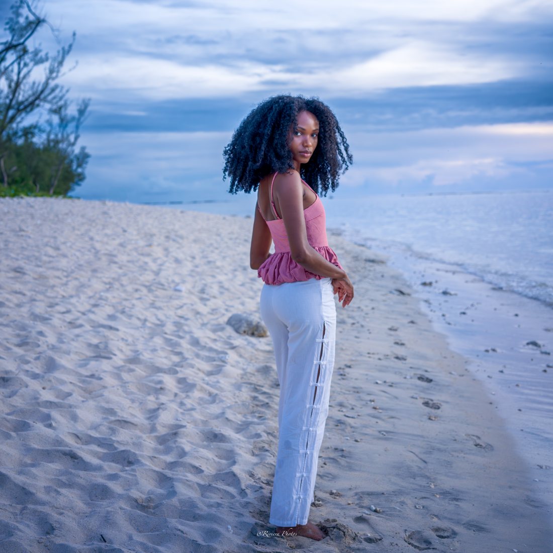 Jeune femme en haut rose à petits carreaux et pantalon blanc, pieds nus sur le sable clair au bord du lagon, filaos à gauche, lumière bleutée de fin de journée.