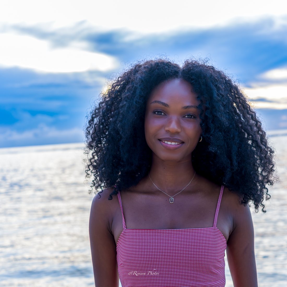 Portrait souriant d'une jeune femme aux cheveux bouclés, haut rose vichy, le lagon et le ciel bleu en fond.