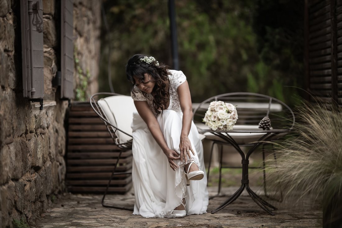 Pendant les préparatifs, la mariée en robe blanche attache sa chaussure, assise sur une terrasse de pierre, son bouquet de roses posé à côté d'elle.