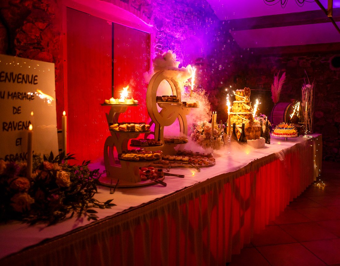 La salle de réception le soir, une longue table de desserts et de bougies sous une lumière rouge et violette, un panneau de bienvenue au premier plan.