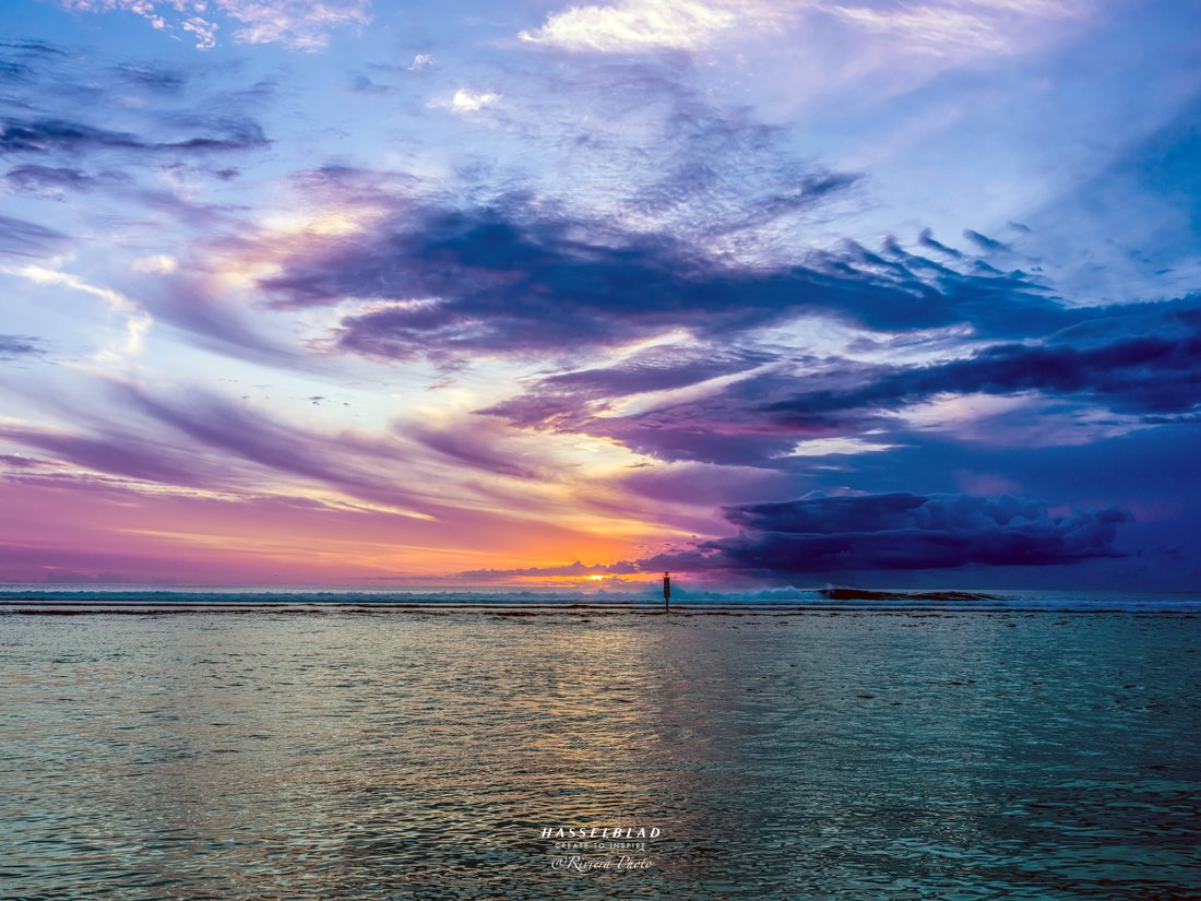 Coucher de soleil violet et orange sur un lagon, une balise dans le lagon au loin.